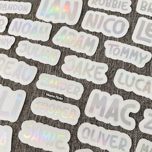 May include: A collection of die-cut stickers featuring various names such as Ian, Noah, Adam, Nico, Tommy, Julian, Jake, Beau, Zander, Lucas, Cooper, Mac, Jamie, and Oliver. Each sticker has a matte holographic finish with white borders.