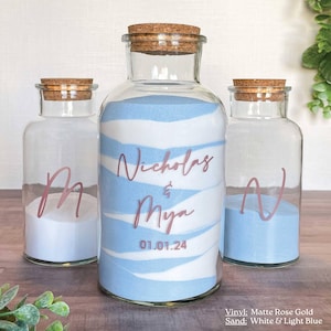 May include: Three clear glass bottles with cork stoppers. The center bottle has white and light blue sand, with the names "Nicholas & Mya" and the date "01.01.24" in matte rose gold vinyl. The other bottles have white and light blue sand and the letters "M" and "N".