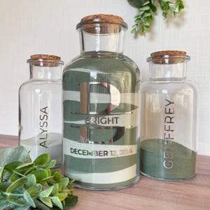 May include: Three clear glass jars with cork stoppers filled with sage green sand. The center jar features a large silver letter B, the name BRIGHT, and the date DECEMBER 12, 2024. The side jars are engraved with the names ALYSSA and GEOFFREY.