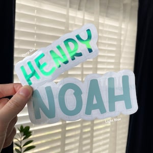May include: Two die-cut vinyl stickers with white borders. The top sticker features the name HENRY in a metallic green finish. The bottom sticker features the name NOAH in a matte light blue finish. Both stickers have a rounded, bubbly font.