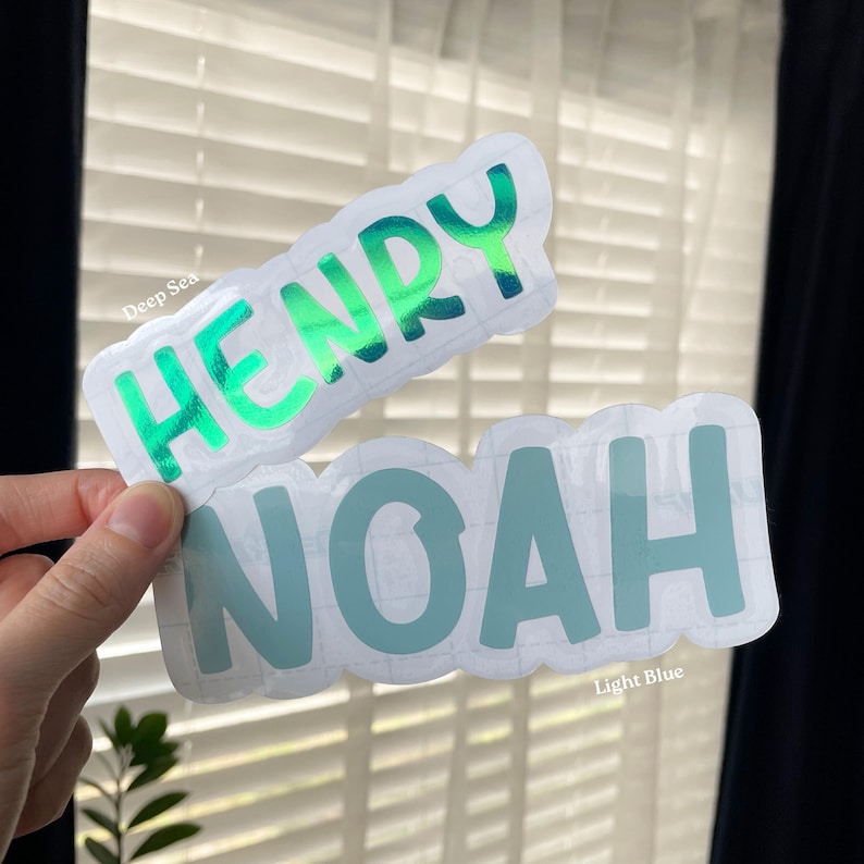 May include: Two die-cut vinyl stickers with white borders. The top sticker features the name HENRY in a metallic green finish. The bottom sticker features the name NOAH in a matte light blue finish. Both stickers have a rounded, bubbly font.