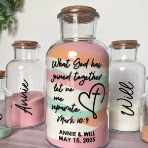 May include: A set of glass bottles with cork stoppers, filled with colored sand. The largest bottle has text that reads "What God has joined together let no one separate. Mark 10:9 ANNIE & WILL MAY 15, 2025". Smaller bottles are labeled with names.