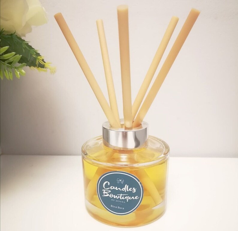 K&ouml;nnte beinhalten: Eine klare Glasdiffuserflasche mit silbernem Deckel und nat&uuml;rlichen Schilfrohrst&auml;ben. Die Flasche enth&auml;lt eine gelbe Fl&uuml;ssigkeit und tr&auml;gt ein Etikett mit der Aufschrift "Candles Boutique by Donna Bora Bora".