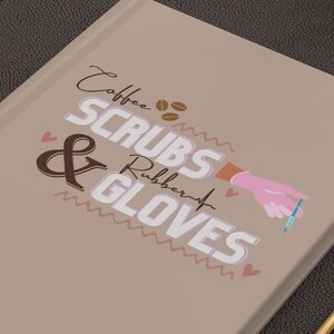 Peut inclure: Un carnet beige avec le texte "Coffee Scrubs & Rubber Gloves" en lettres blanches et roses. Le carnet a une main rose avec une aiguille bleue sur la couverture.