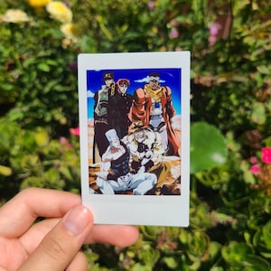 Może przedstawiać: Biały wydruk w stylu polaroid z grupą postaci z anime w pustynnym krajobrazie. Postacie pozują i noszą różne stroje. Wydruk jest trzymany na tle zielonych liści.
