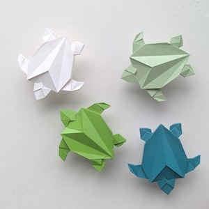May include: Four origami turtles in white, green, light green, and teal blue. The paper turtles are arranged on a white surface. The turtles have a geometric, angular design, with folded paper creating the shape.