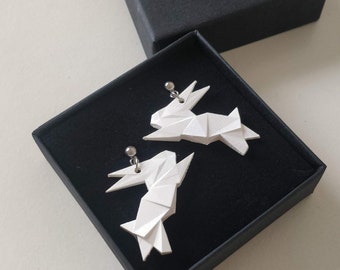 Può includere: Un paio di orecchini a forma di coniglio origami bianco in una scatola nera con il logo "PAINT IT WILD" sul coperchio.