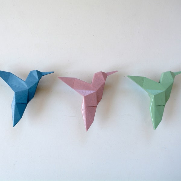 Op de afbeelding: Vier origami-kolibries in verschillende kleuren: wit, blauw, roze en groen. De kolibries zijn gemaakt van papier en hebben een geometrisch ontwerp.