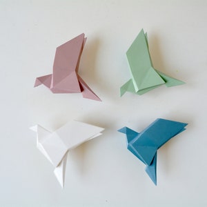 Peut inclure: Quatre oiseaux en papier origami de différentes couleurs : rose, blanc, vert et bleu. Les oiseaux sont pliés dans un style origami traditionnel et sont disposés en carré sur un fond blanc.