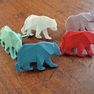 以下が含まれることがあります： 白、緑、青、赤、紫の5色の幾何学的なクマの形をした引き出しの取っ手。取っ手は滑らかで硬い素材で作られており、モダンでミニマルなデザインです。