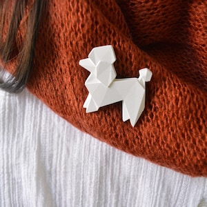 May include: A white, geometric poodle brooch. The brooch is affixed to a rust-colored scarf and a white textured shirt. The poodle design is angular and modern, with a minimalist aesthetic. The brooch adds a touch of whimsy to the outfit.