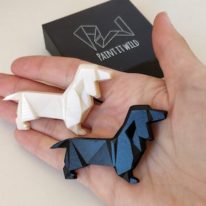 May include: Two origami dachshund figurines, one white and one black, are held in a hand. The figurines are made of paper and have a geometric design. The background includes a black box with the text "Paint It Wild".