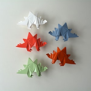 Puede incluir: Cinco dinosaurios de origami en diferentes colores: blanco, rojo, azul, naranja y verde. Los dinosaurios están todos mirando en la misma dirección y tienen una espalda espinosa.