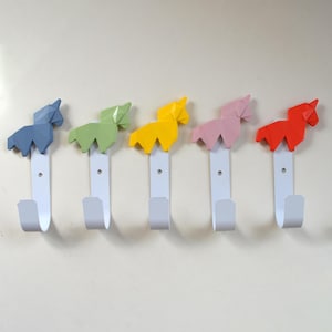 Könnte beinhalten: Fünf Wandhaken mit weißer Basis und farbenfrohem Origami-Pferdedesign. Die Pferde sind blau, grün, gelb, rosa und rot. Die Haken sind an einer hellfarbigen Wand befestigt.