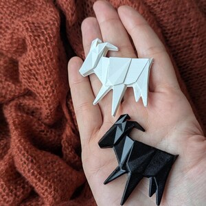 Könnte beinhalten: Zwei Origami-Ziegen, eine weiße und eine schwarze, werden in der Hand einer Person gehalten. Die Ziegen sind aus Papier und haben ein geometrisches Design.