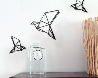 Puede incluir: Tres decoraciones de pared de pájaros origami de metal negro, un reloj de vidrio transparente con números romanos y un jarrón lleno de ramas de madera flotante sobre una superficie de madera oscura.