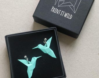 Può includere: Un paio di orecchini a forma di colibrì origami blu turchese in una scatola nera con la scritta "PAINT IT WILD" sul coperchio.