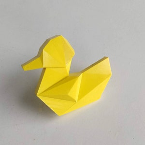 May include: A yellow geometric duck figurine made of plastic. The duck has a simple design with a triangular beak and a rounded head.
