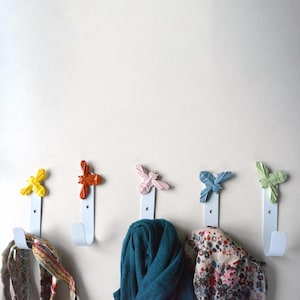 Può includere: Cinque ganci in metallo bianco con un design a forma di ape in diversi colori. I ganci sono montati su una parete bianca. Una sciarpa blu, una sciarpa rosa e bianca e una sciarpa gialla e marrone sono appese ai ganci.