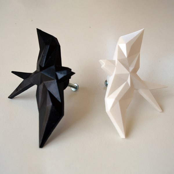 Op de afbeelding: Twee geometrische vogelvormige kastknoppen, één zwart, de andere wit. De knoppen zijn gemonteerd op zilveren schroeven en hebben een modern, hoekig uiterlijk. Deze decoratieve knoppen zijn geschikt voor laden of kasten.