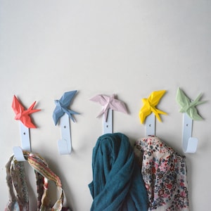 Op de afbeelding: Vijf witte metalen wandhaken in de vorm van origami-vogels in verschillende kleuren: rood, blauw, roze, geel en groen. De haken zijn bevestigd aan een witte muur en er hangen sjaals aan.