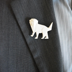Peut inclure: Une broche en origami en forme de chat, de couleur blanche, fixée sur une veste de costume gris foncé.
