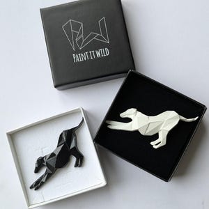 Op de afbeelding: Twee zwarte dozen met witte binnenkant, elk met een geometrische, gestileerde zwarte en witte hondenbroche. De dozen hebben het logo "PAINT IT WILD" op de bovenkant gedrukt.