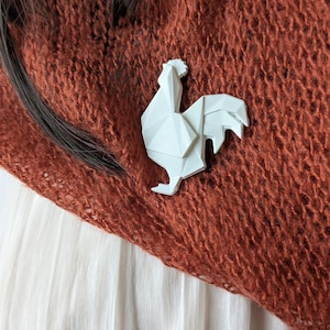 May include: White origami rooster brooch pinned to a rust-colored knitted scarf.