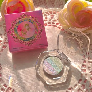 May include: A pink and gold compact with a unicorn design and the words "Rainbow Eyeshadow" and "Net Wt. 3.00g". The compact is open, revealing a clear container with a light blue eyeshadow with iridescent glitter.