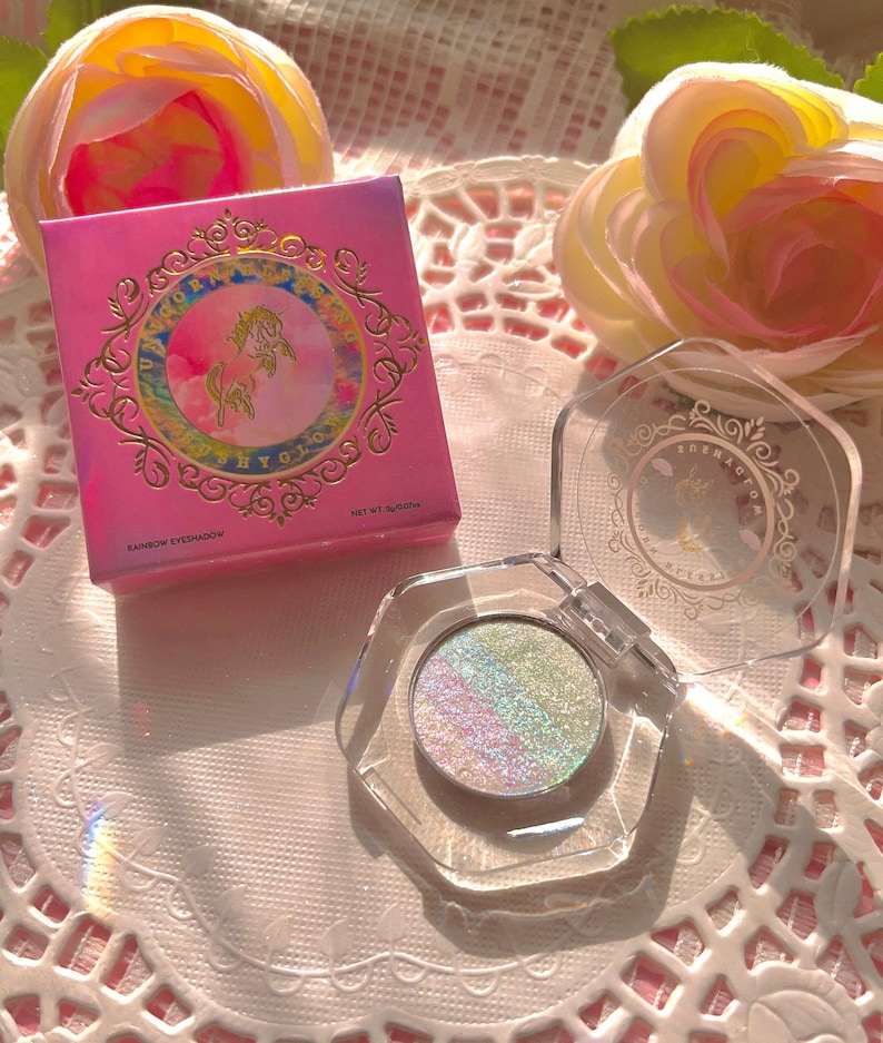 May include: A pink and gold compact with a unicorn design and the words "Rainbow Eyeshadow" and "Net Wt. 3.00g". The compact is open, revealing a clear container with a light blue eyeshadow with iridescent glitter.