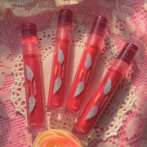 Pode incluir: Quatro tubos de gloss labial rosa com um design de asa de anjo branco e o texto "Sushi Glow Cosmetics" e "Fantasy Lipgloss". Cada tubo contém 7 ml de gloss labial.