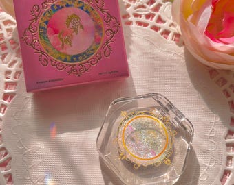 May include: A pink and gold compact with a unicorn design and the text "Unicorn Dreaming" and "Mushy Glow" on the front. The compact contains a clear, iridescent eyeshadow with glitter.