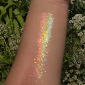 May include: A close-up of iridescent glitter on skin, showing a rainbow of colors, including pink, yellow, green, and blue.