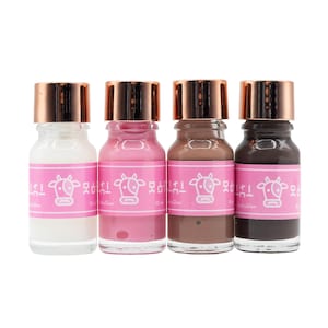 Pode incluir: Quatro pequenos frascos de vidro com tampas de cobre metálico, cada um contendo 10 ml de líquido nas cores branca, rosa, castanha e preta. Cada frasco tem um rótulo rosa com uma ilustração de uma vaca branca e o texto "MooGlow".