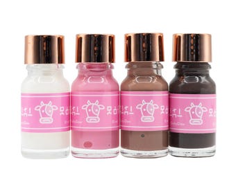 Pode incluir: Quatro pequenos frascos de vidro com tampas de cobre metálico, cada um contendo 10 ml de líquido nas cores branca, rosa, castanha e preta. Cada frasco tem um rótulo rosa com uma ilustração de uma vaca branca e o texto "MooGlow".