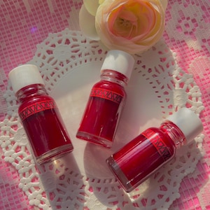 May include: Three small glass bottles with white caps filled with a deep red liquid. The bottles are sitting on a white doily with a delicate lace pattern. The doily is on a pink and white checkered background.