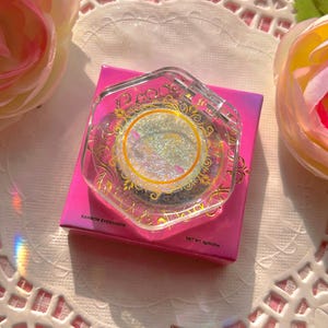 May include: A single round iridescent rainbow eyeshadow in a clear plastic container with a gold design. The container is sitting on a pink and white background. The text on the container reads "Rainbow Eyeshadow" and "Net Wt. 5g/0.17oz".