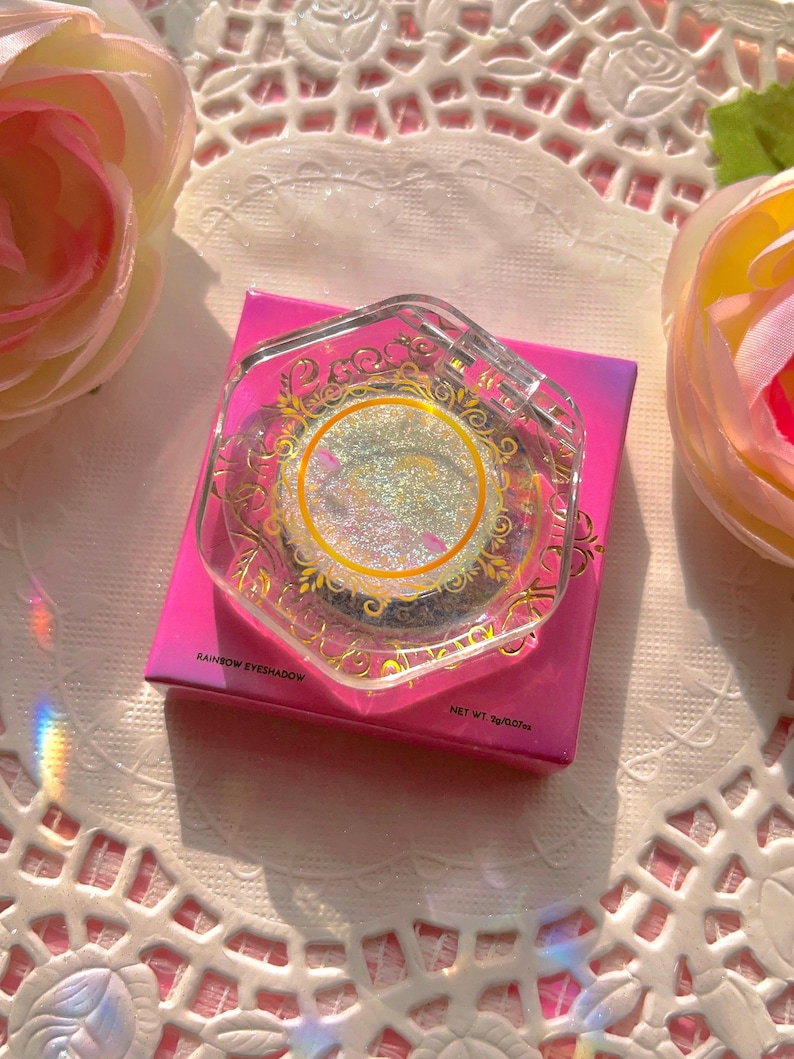 May include: A single round iridescent rainbow eyeshadow in a clear plastic container with a gold design. The container is sitting on a pink and white background. The text on the container reads "Rainbow Eyeshadow" and "Net Wt. 5g/0.17oz".