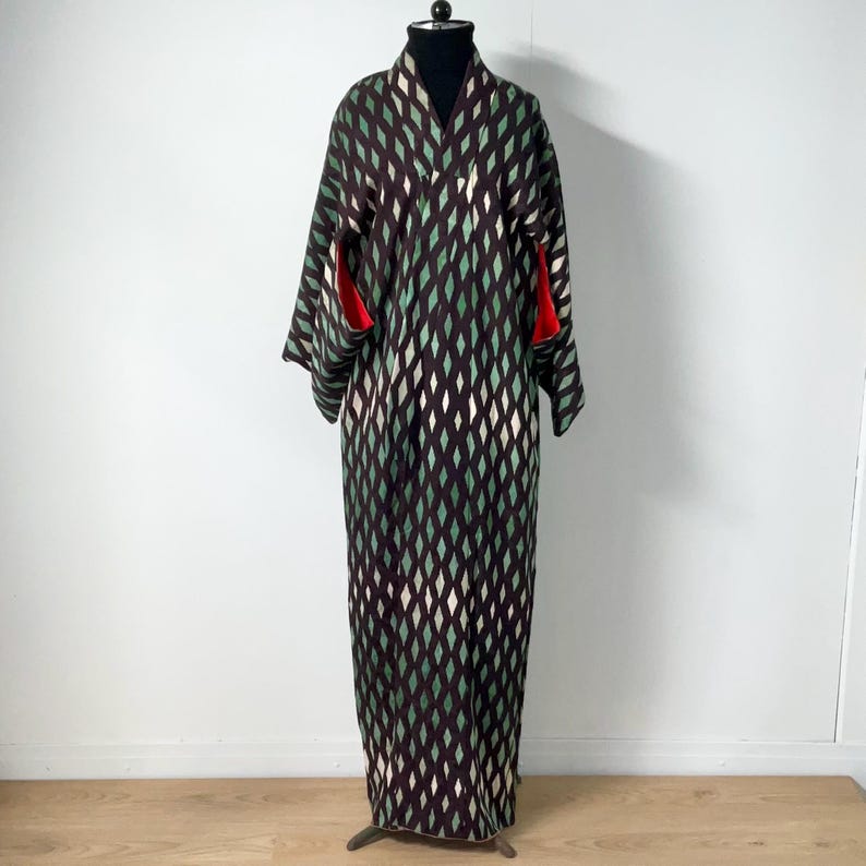 May include: A full-length kimono with a diamond pattern in shades of brown, green, and white. The kimono has long sleeves with red accents at the cuffs. It is displayed on a black mannequin head and stand.