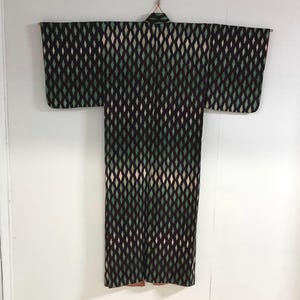 May include: A vintage kimono with a repeating diamond pattern in black, green, and cream. The kimono is hanging against a white background, showcasing its full length and wide sleeves. The fabric appears to be a woven textile.