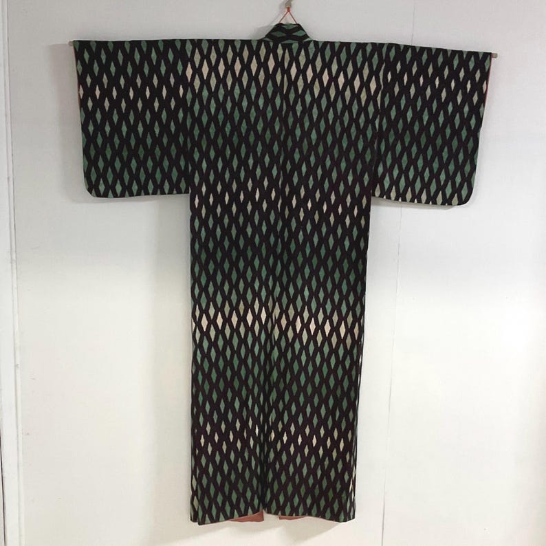May include: A vintage kimono with a repeating diamond pattern in black, green, and cream. The kimono is hanging against a white background, showcasing its full length and wide sleeves. The fabric appears to be a woven textile.