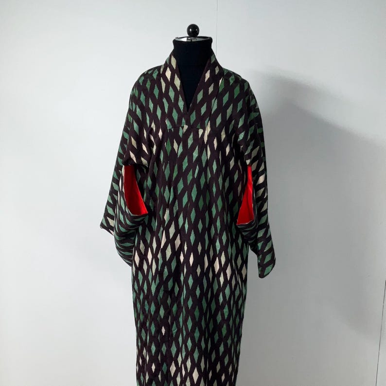 May include: A long, patterned kimono in shades of black, green, and white, displayed on a mannequin. The kimono features a diamond pattern and red accents on the sleeves. The garment is made of a soft, flowing fabric.