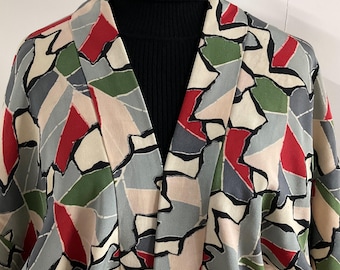 May include: A patterned kimono-style jacket with a geometric design in red, green, gray, and beige. The jacket has a V-neck and is worn over a black turtleneck. The pattern is outlined in black.