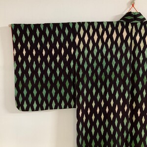 May include: A black kimono with a repeating diamond pattern in shades of green and off-white. The kimono is hanging against a white background, showcasing the intricate design and the garment's structure.