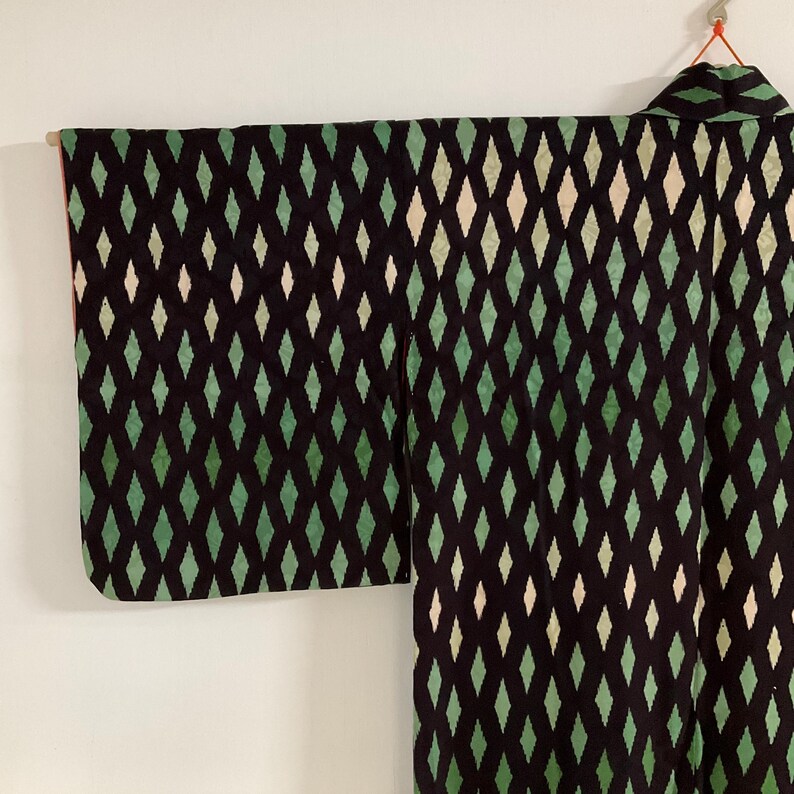May include: A black kimono with a repeating diamond pattern in shades of green and off-white. The kimono is hanging against a white background, showcasing the intricate design and the garment's structure.