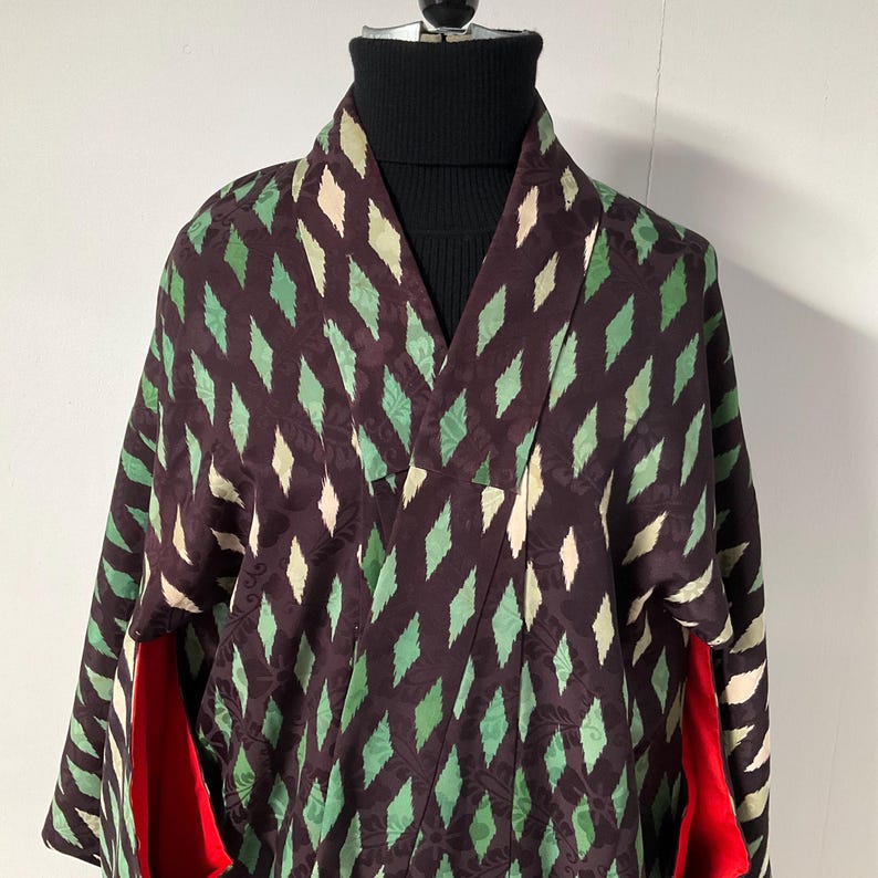May include: A dark brown kimono with a repeating diamond pattern in green and off-white. The kimono has a wide collar and long sleeves. The interior lining of the sleeves is red. The kimono is displayed on a black turtleneck.