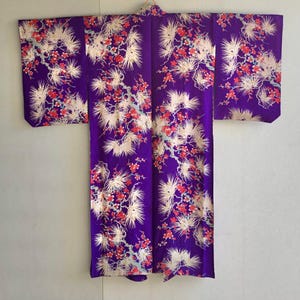 May include: A vibrant purple kimono with a floral pattern. The design features white, red, and gold-colored blossoms and foliage. The garment is displayed flat, showcasing its full length and intricate details.