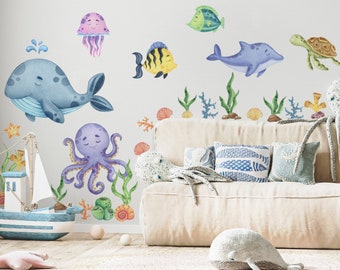 May include: A set of colorful watercolor-style ocean animal wall decals featuring a whale, octopus, jellyfish, fish, turtle, and seaweed. The decals are shown on a white wall above a light-colored couch with pillows.