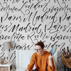 Puede incluir: Una persona se sienta en una cama con un edredón blanco y una pared blanca con nombres de ciudades escritos en negro en cursiva, incluyendo París, Londres, Madrid y Barcelona. La persona lleva un jersey naranja brillante y unos vaqueros azules. Está mirando un ordenador portátil en la cama.