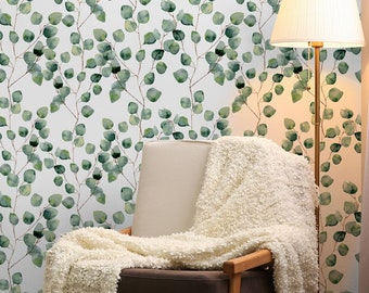 May include: A white wall with a green leafy pattern wallpaper. A white armchair with a white throw blanket is in the corner of the room. A gold floor lamp with a white lampshade is next to the chair. A wooden shelf with a teddy bear and pillows is in the foreground.